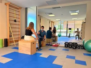 Sala de rehabilitación con piso azul, cajones de madera y pesas rusas, con personas conversando al fondo