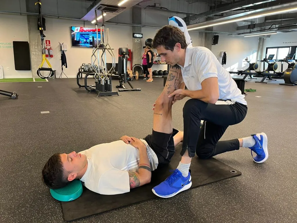 Fisioterapeuta movilizando la pierna de un paciente acostado sobre una colchoneta en un gimnasio amplio