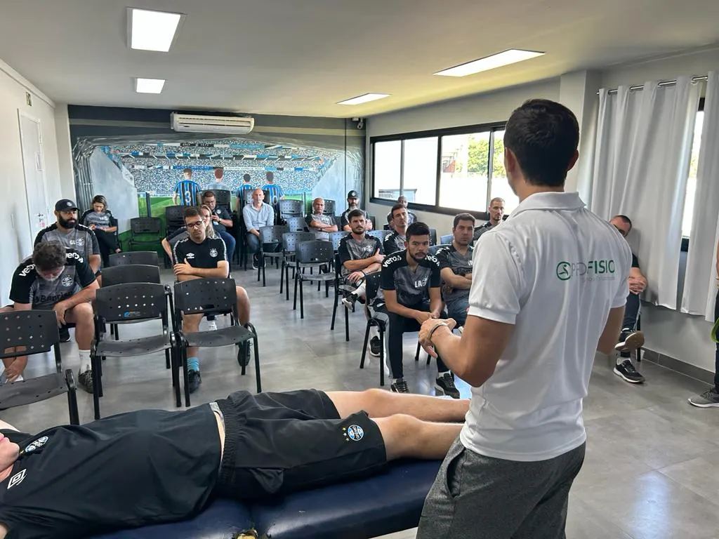 Profesional dando una charla a un grupo de deportistas sentados, con una persona acostada en una camilla